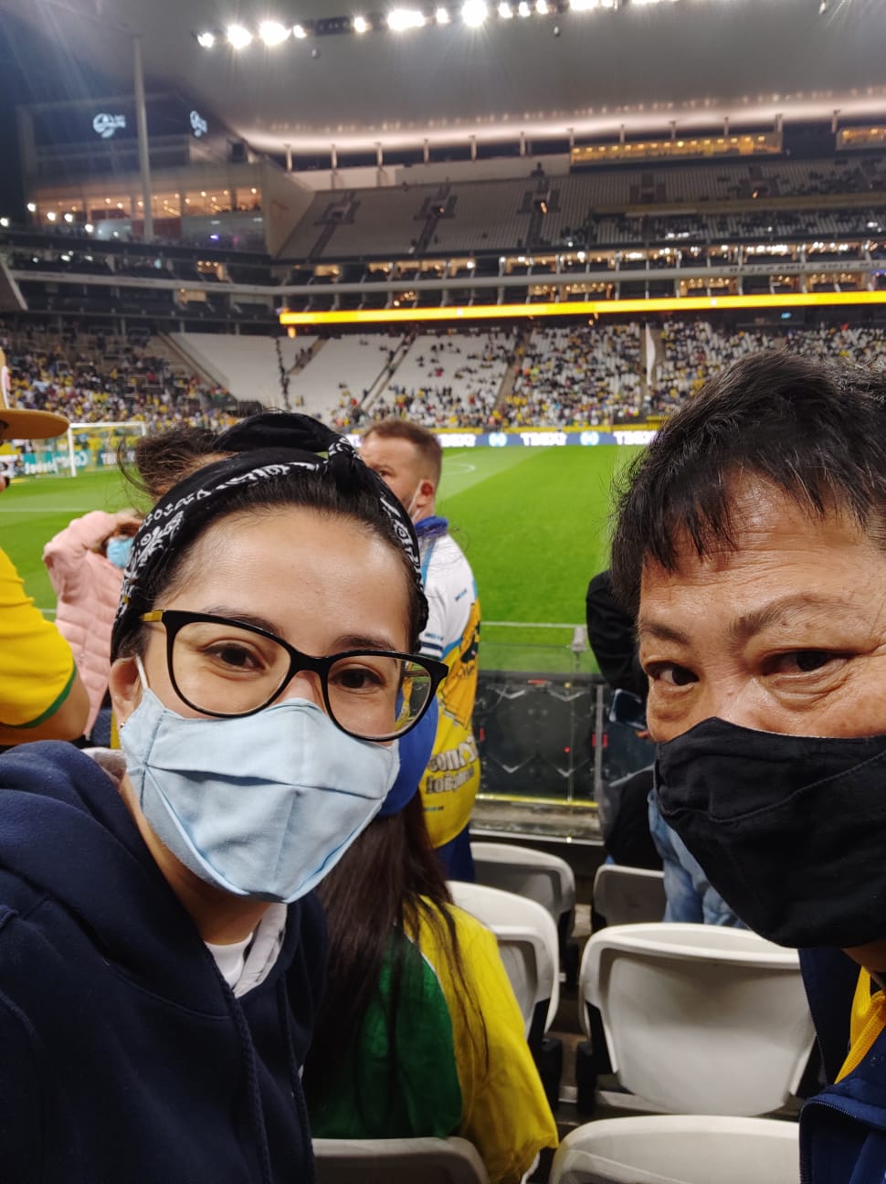 Uma mulher usando óculos e lenço amarrado no cabelo está ao lado de um homem. Os dois estão usando máscara de proteção. Atrás deles, está um campo de futebol.