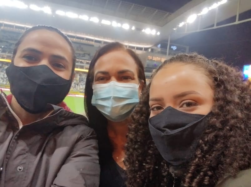 Três mulheres estão usando máscara de proteção na arquibancada de um estádio. Atrás delas, está uma parte do campo de futebol.