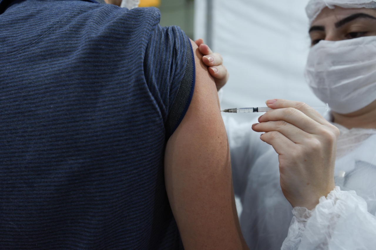 À direita da imagem, uma profissional de saúde vestindo jaleco branco, máscara de proteção branca e touca nos cabelos está aplicando a dose da vacina no braço de uma pessoa, que não tem o rosto mostrado. A pessoa está de costas e veste uma blusa azul com listras preta.