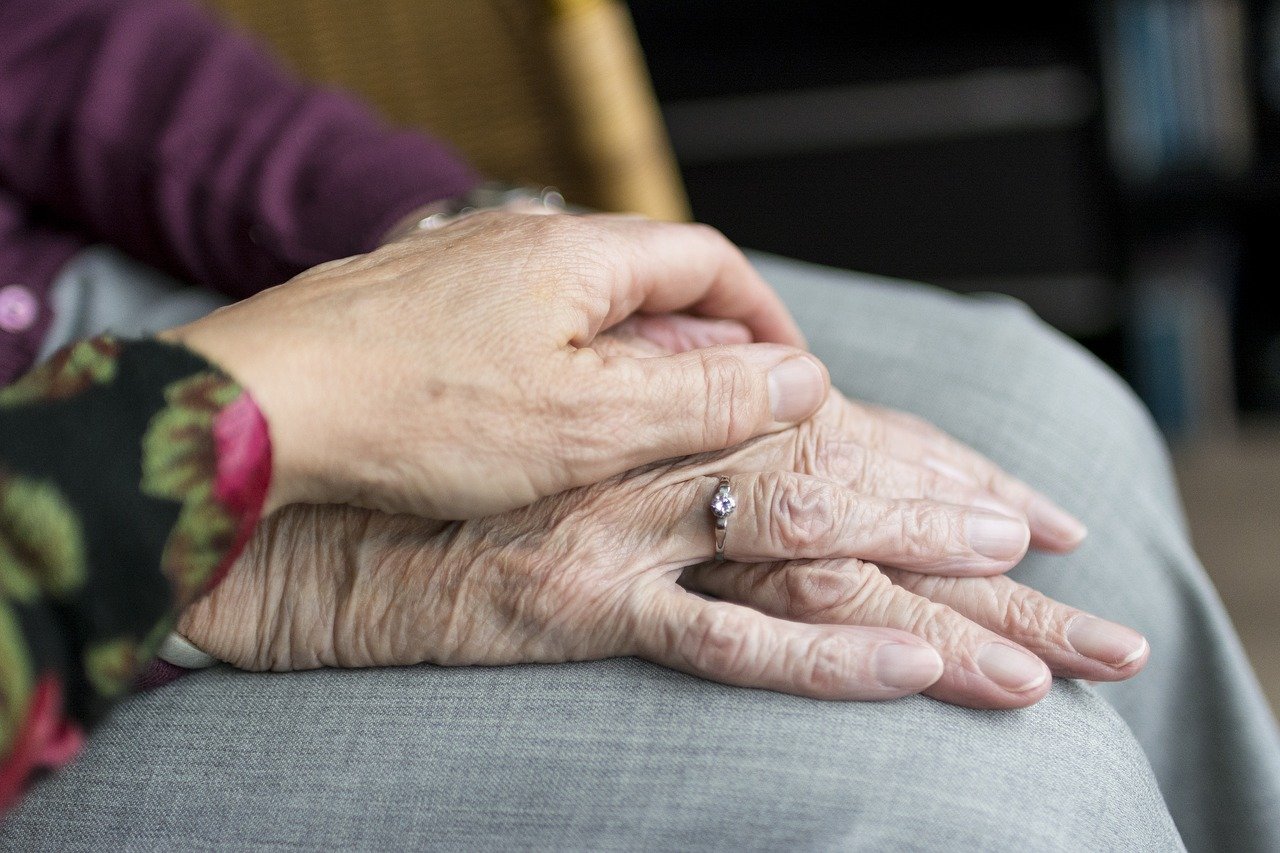 Imagem de uma mão enrugada apoiada em outra mão de um idoso, que está em cima de uma calça cinza. A mão que está embaixo tem um anel fino no dedo anelar.
