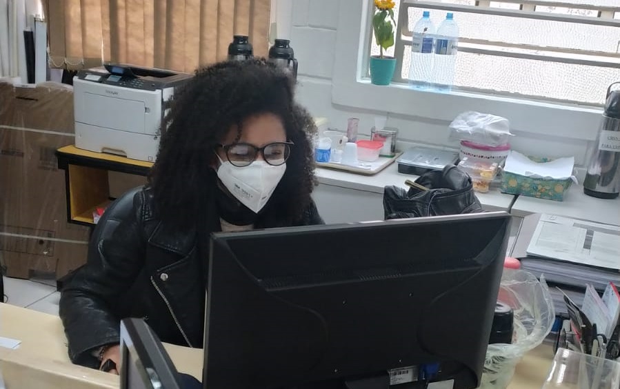 Uma jovem negra com máscara de proteção branca e uma jaqueta de couro está sentada em frente a um computador. Ela usa óculos de grau e está olhando fixamente para a tela. Atrás dela, há uma parede com duas janelas, uma impressora branca apoiada em uma mesa amarela e itens de decoração.