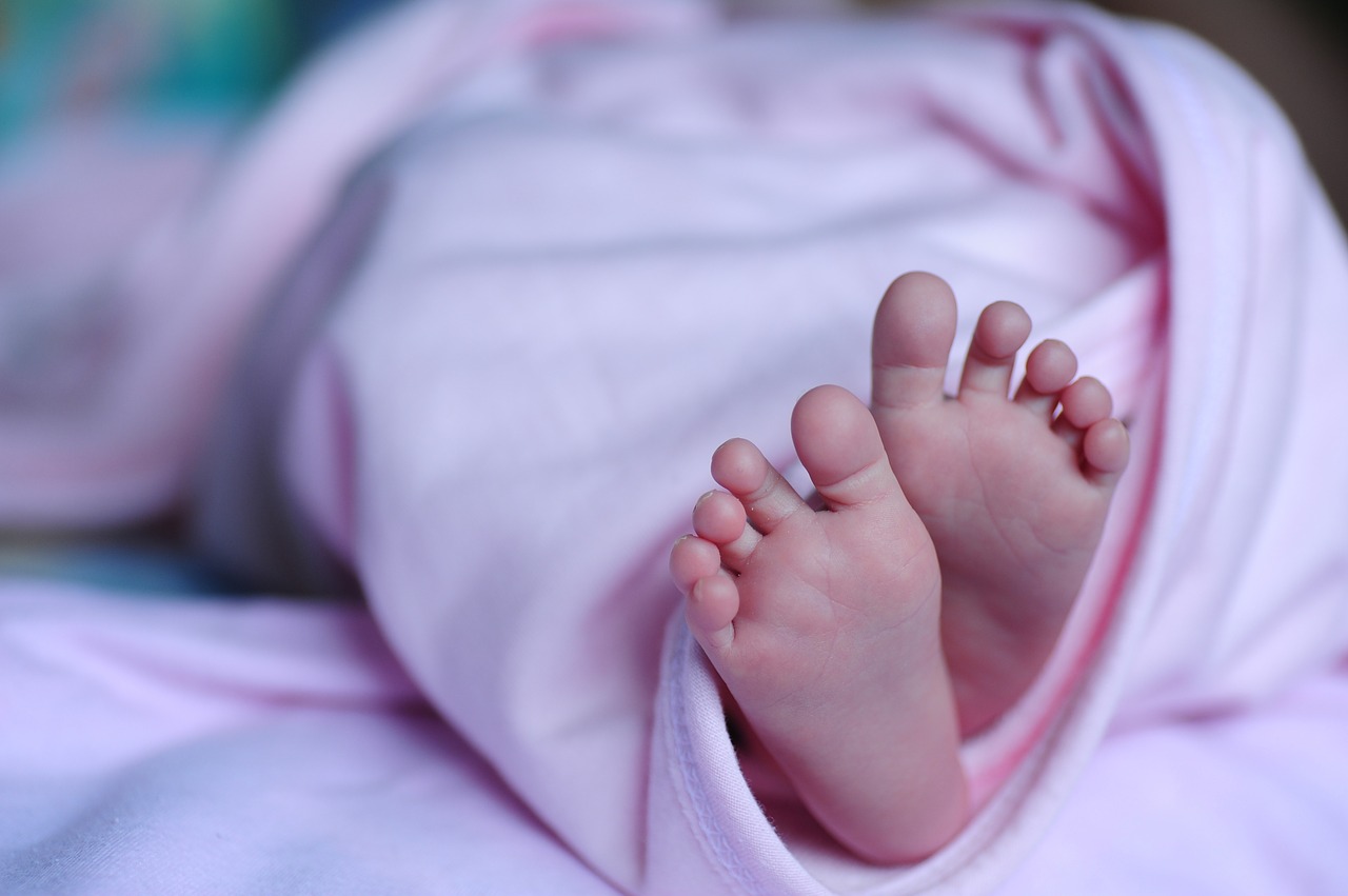 Na imagem, há dois pés de um recém-nascido com o fundo desfocado. Ele está coberto em uma manta rosa.