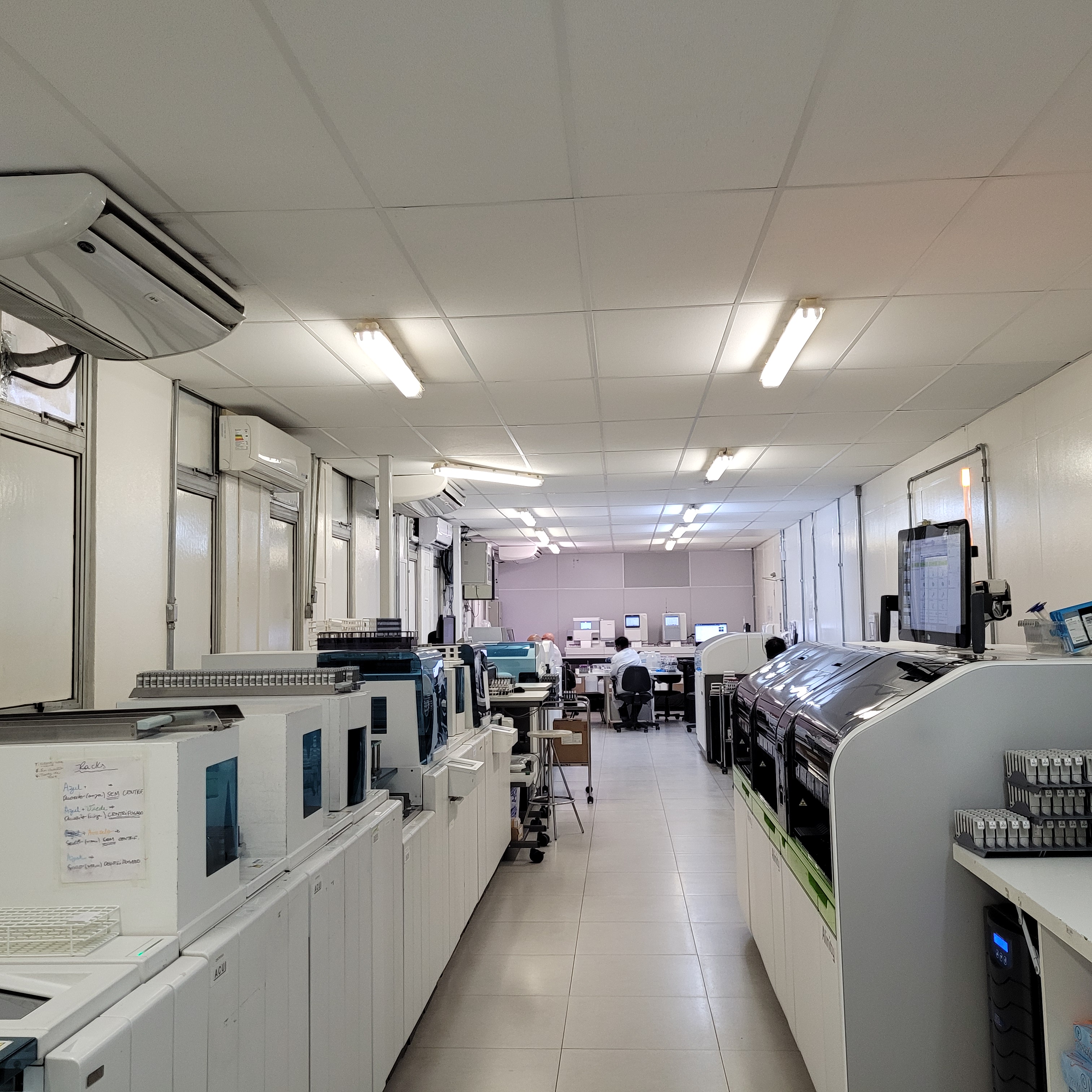 Imagem de uma sala com equipamentos do laboratório. Ao fundo é possível observar as costas de uma profissional sentada à mesa.