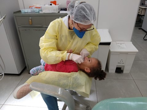 Na imagem, uma profissional de saúde vestindo avental amarelo está segurando uma menina de aproximadamente 3 anos no colo. Ela está com as mãos, cobertas por luvas brancas, na boca da criança, examinando seus dentes.