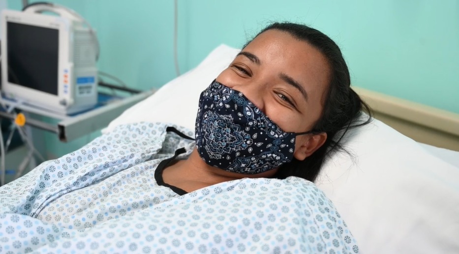 Uma mulher está deitada em uma cama de hospital. Ela veste um avental branco e azul, tem os cabelos presos e usa uma máscara de proteção no rosto.