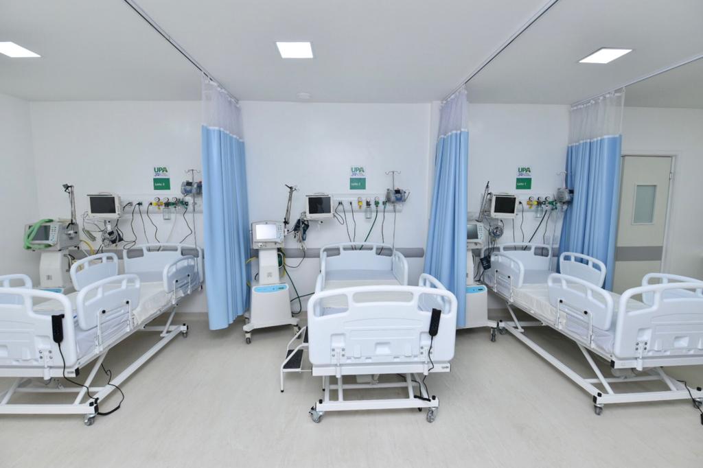 Em uma sala com paredes brancas estão três leitos de hospital vazios, separados por uma cortina azul clara. O cômodo tem equipamentos de internação ao lado das camas.