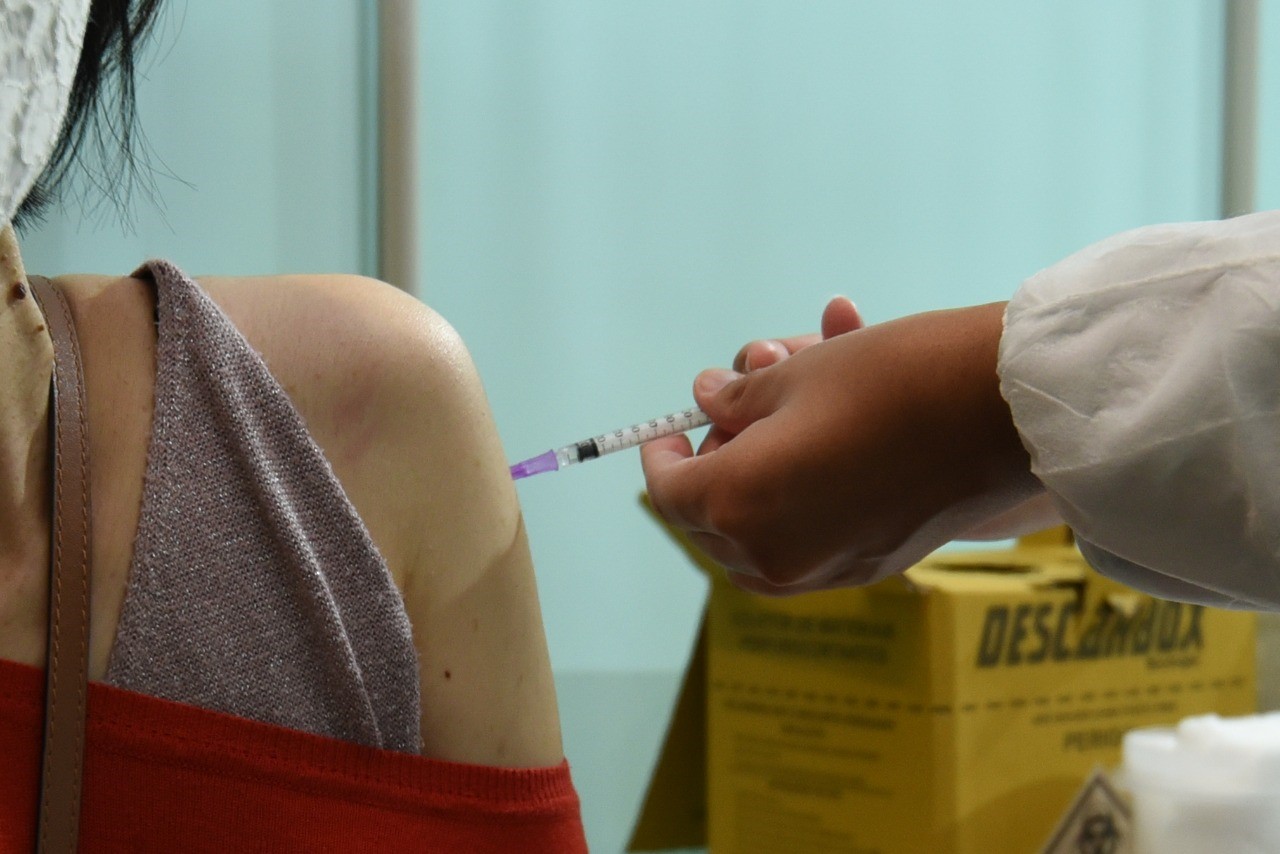 Foto de vacinação. A mão de um profissional de saúde está aplicando a vacina em um braço de uma mulher que veste uma blusa vermelha e usa uma máscara de proteção branca.