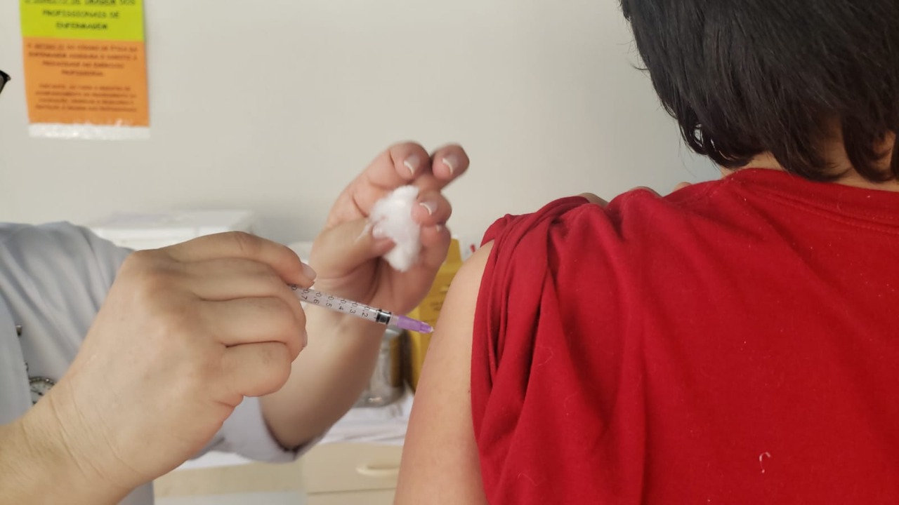 Foto de vacinação. Uma profissional de saúde está aplicando a dose de vacina em um jovem de camisa vermelha, que está de costas e tem cabelos pretos no tamanho médio. A profissional segura a seringa em uma mão e um algodão em outra.
