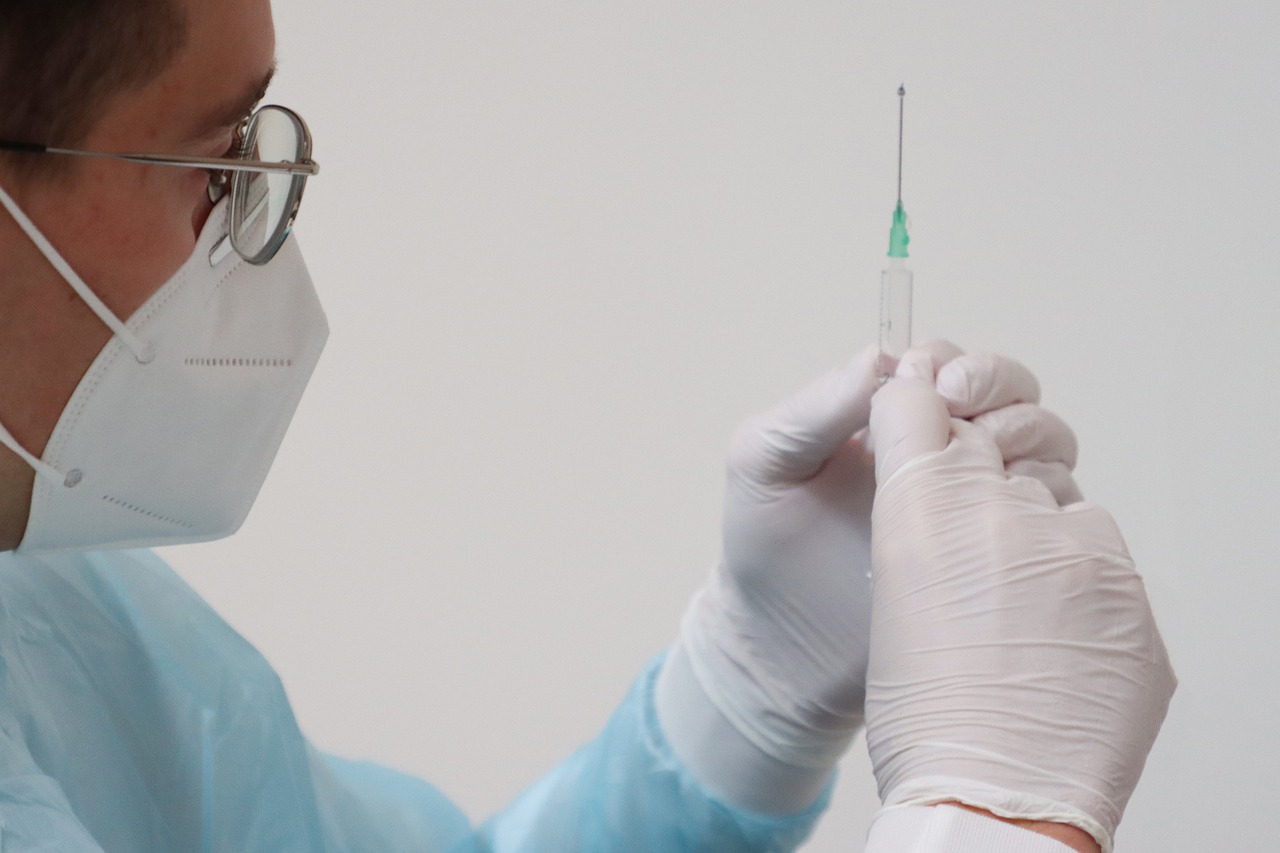 Foto de vacina. Um profissional de saúde vestindo máscara branca e usando óculos de grau está segurando uma agulha, prestes a vacinar alguém. Ele usa luvas brancas.