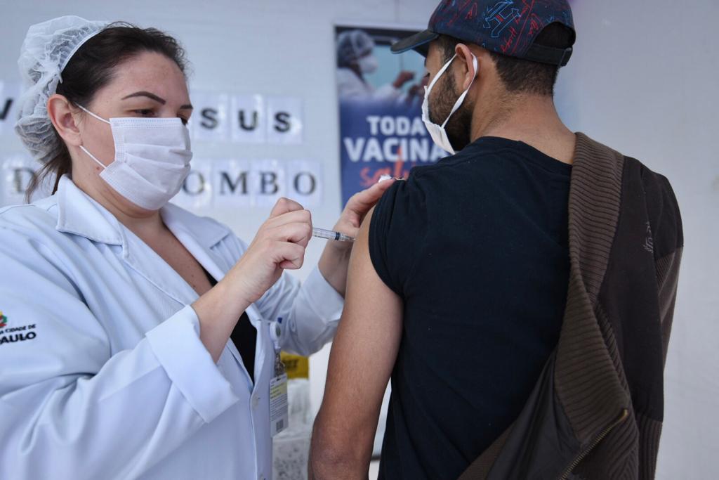 Uma profissional de saúde vestindo jaleco branco está aplicando a dose da vacina no braço de um rapaz mais alto, que está ao seu lado. Ele está de costas, usa uma camisa preta que está com uma das mangas levantadas e um boné azul e vermelho.