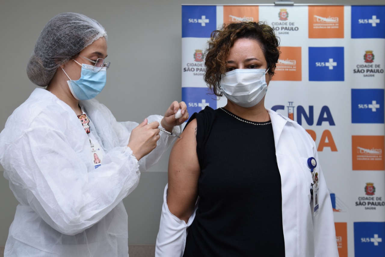 Foto de vacinação. À direita da imagem, uma mulher de cabelos curtos cacheados está recebendo a vacina, aplicada por uma profissional de saúde que está à esquerda da foto. A mulher tem o jaleco branco abaixado no braço que está recebendo a dose.