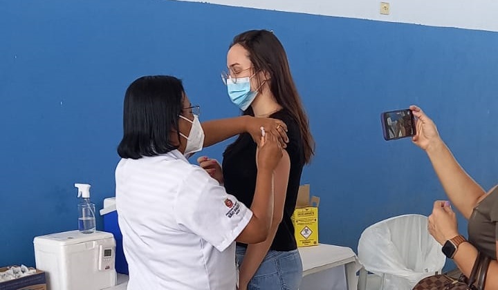 Foto de vacinação. Uma profissional de saúde vestindo um jaleco branco está aplicando a vacina em uma jovem de camisa preta e calça jeans clara. Ao fundo, está uma parede azul. À direita da imagem, uma pessoa tira uma foto do momento com um celular. 
