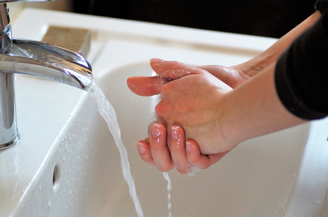 Duas mãos femininas com as unhas levemente grandes, sem esmalte, estão em frente a uma torneira aberta, com água descendo. Embaixo, há uma pia branca.