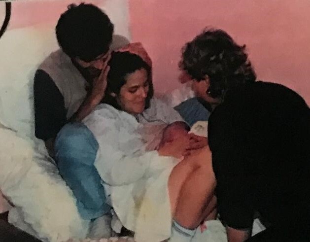 Um homem está sentado atrás de uma mulher vestida de branco, que está segurando um recém-nascido no colo. Em frente a ela, está uma mulher virada de costas, olhando para o bebê.