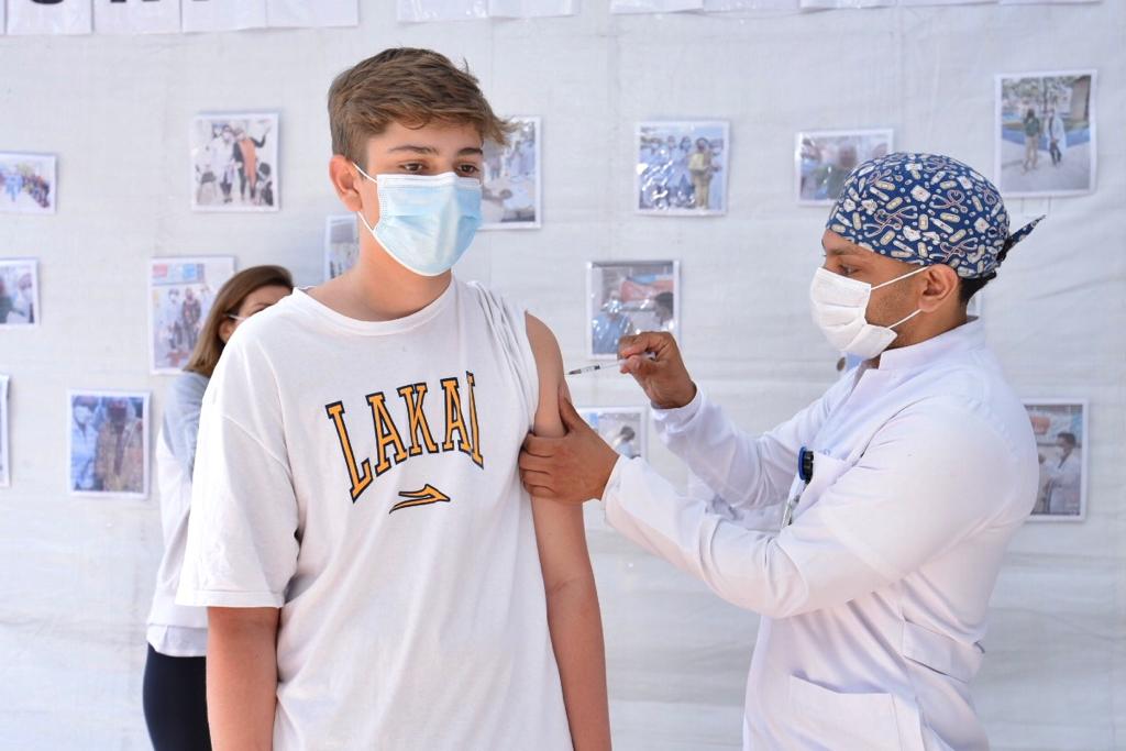 Um profissional de saúde vestindo jaleco branco, máscara de proteção branca e uma touca azul com detalhes em branco na cabeça está aplicando a dose da vacina em um jovem mais alto. Ele veste uma camisa branca com detalhes em amarelo.