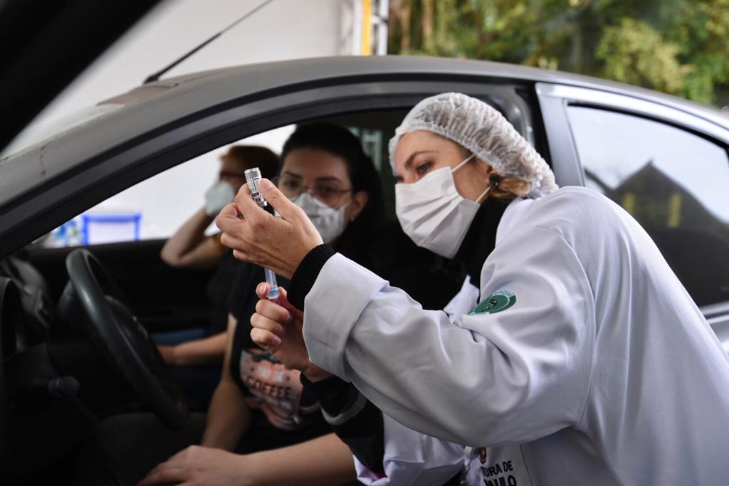 Foto de vacinação. Uma profissional de saúde usando jaleco branco, máscara de proteção branca e touca nos cabelos está em frente a um carro preto com a porta aberta, mostrando o frasco da vacina para uma mulher, que está sentada no banco do motorista do carro.