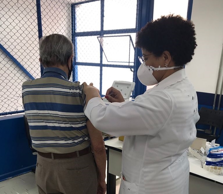 Foto de vacinação. Uma profissional da saúde usando jaleco, máscara e óculos de grau está aplicando vacina em homem idoso que está de costas vestindo camisa com listras azuis, brancas e beges. Ao fundo há uma janela aberta.