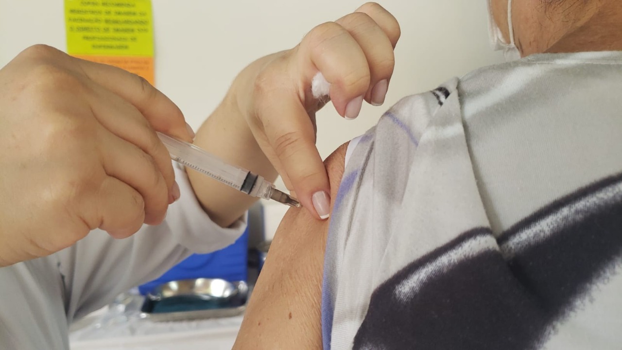 Foto de vacinação. Enquadramento da foto está fechado em aplicação da vacina. Profissional da saúde aplica vacina em braço de idosa usando roupa cinza com listra azul com a manga levantada.