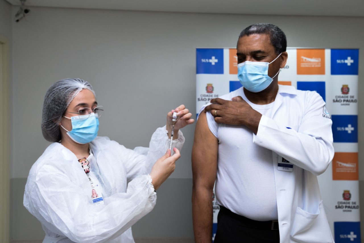 Foto de vacinação. À direita da imagem, um homem está recebendo a dose da vacina, aplicada por uma profissional de saúde, que está à esquerda da imagem. Ele tem o jaleco branco pendurado em um ombro.