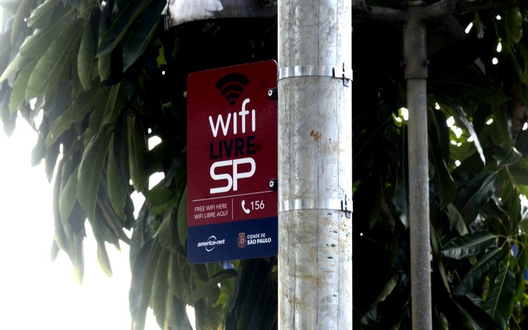 foto de poste com placa fixada, na placa há o símbolo do wifi, logo abaixo o texto" Wifi livre SP", abaixo do texto ícone de telefone e os números 156, no rodapé sob plano de fundo azul o logotipo da Prefeitura de São Paulo