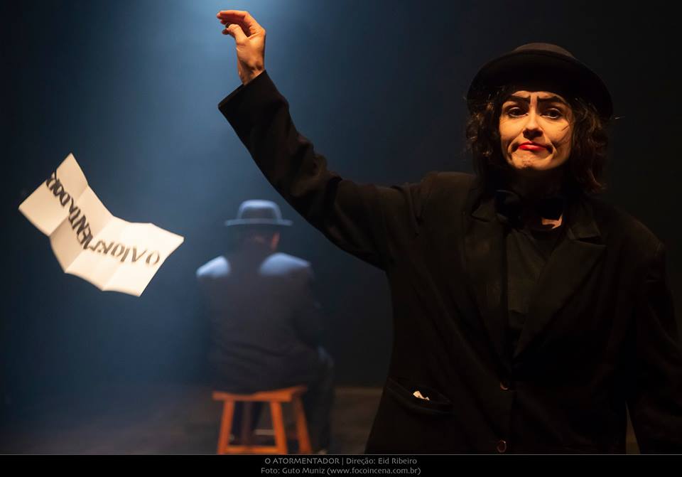 Foto de mulher em palco; ela veste paletó preto e chapéu da mesma cor e solta uma folha da mão direiita