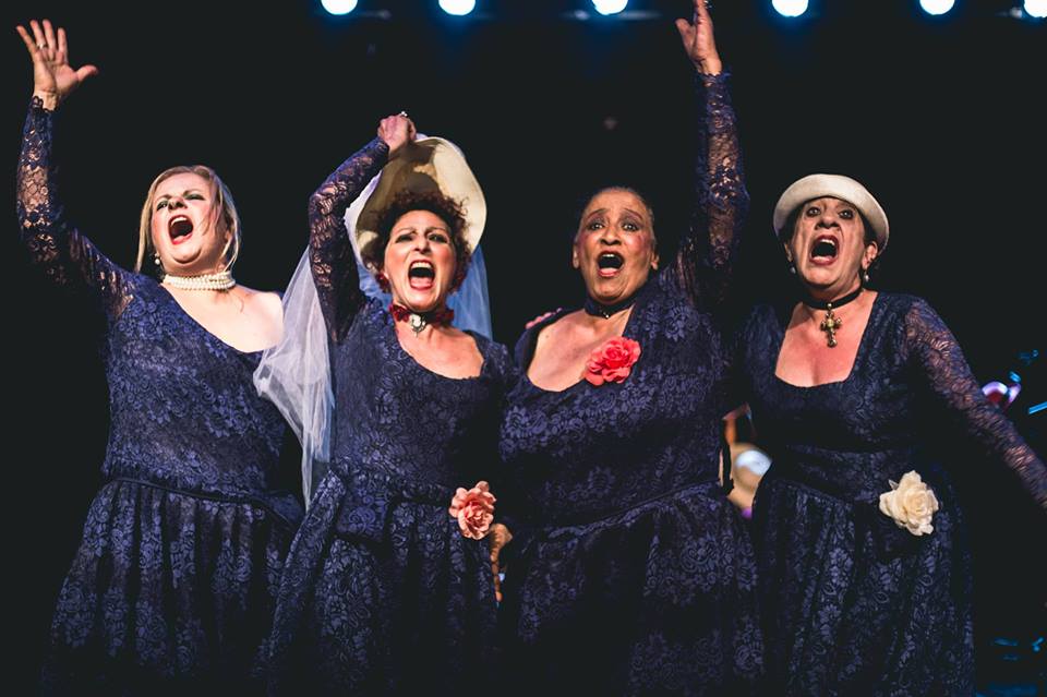 Foto de quatro mulheres com vestidos roxos, mãos para o alto e chapéus. Elas estão com a boca aberta, como se estivessem gritando.