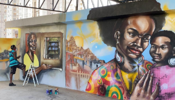 Parede grafitada com desenhos de mulheres negras, artista trabalhando numa delas no canto esquerdo