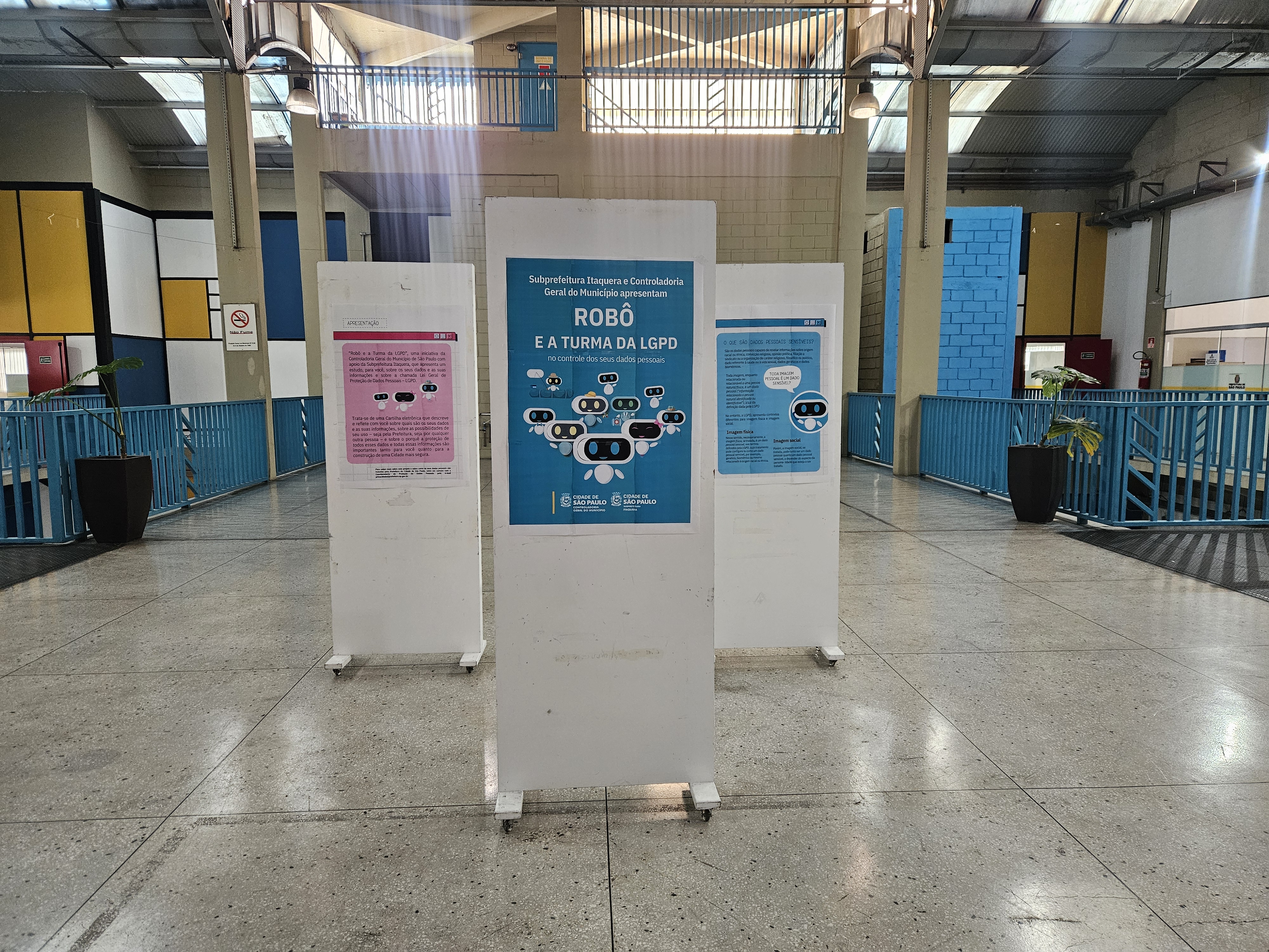 Foto da exposição no salão da Subprefeitura de Itaquera, na imagem tem 3 paineis brancos, cada um com uma página da revista Robê e a Turma da LGPD