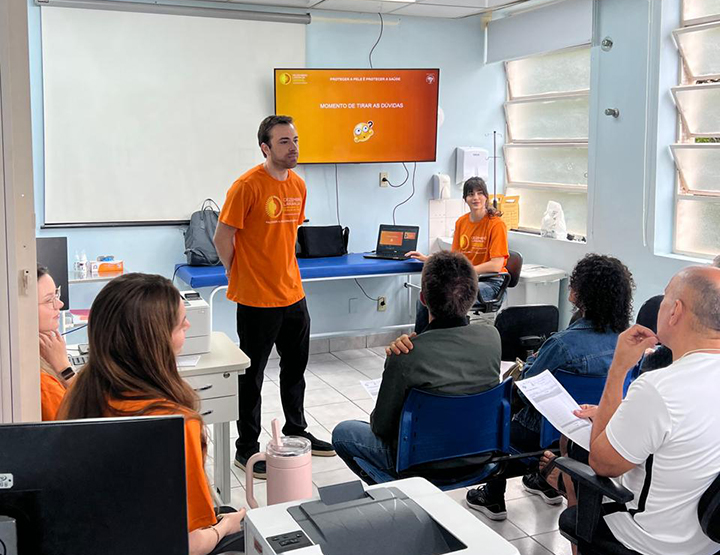 A imagem mostra uma sala com paredes azul-claro e janelas grandes à direita. No centro da cena, um homem em pé, vestindo uma camiseta laranja, fala para um grupo de pessoas sentadas. O homem que está em pé no centro da sala parece ter estatura média e porte físico magro. Ele tem pele clara e cabelos curtos, castanhos, penteados para o lado. Seu rosto é liso, sem barba, e sua expressão é séria e concentrada, transmitindo atenção ao que está explicando. À sua volta, outras duas pessoas também usam camisetas laranja e estão sentadas, observando a apresentação. Ao fundo, há uma mesa com notebook e materiais, e acima dela uma televisão ligada exibindo um slide com o título “Momento de tirar as dúvidas”. As pessoas que assistem estão sentadas em cadeiras azuis, algumas segurando folhas de papel e prestando atenção ao que é explicado. O ambiente passa a sensação de encontro formativo ou instrutivo, com clima de diálogo e participação