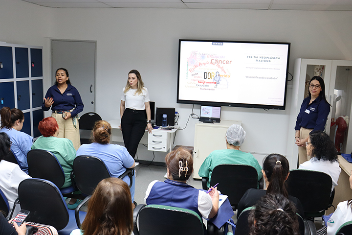 Foto colorida em ambiente interno, uma sala de reunião. Várias pessoas adultas estão sentadas em cadeiras, vistas principalmente de costas, assistindo a uma apresentação. A maioria usa uniformes ou roupas de trabalho da área da saúde, em tons de azul, verde e branco. Na parte da frente da sala, três mulheres estão em pé. Duas delas falam ao grupo e uma observa. Elas vestem camisas sociais e calças, com aparência profissional. Atrás delas, há uma televisão grande fixada na parede exibindo um slide com palavras relacionadas à saúde, como “câncer” e “dor”, em destaque. O ambiente é organizado, com armários à esquerda, equipamentos eletrônicos próximos à tela e iluminação clara. A cena transmite um momento de capacitação para profissionais da saúde.
