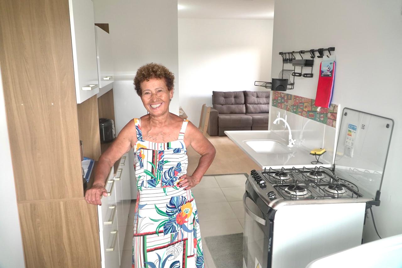 Mulher sorridente, de vestido estampado colorido, está em uma cozinha clara e organizada. Ela apoia uma mão no armário de madeira e a outra na cintura. Ao fundo, dá para ver uma sala com um sofá.