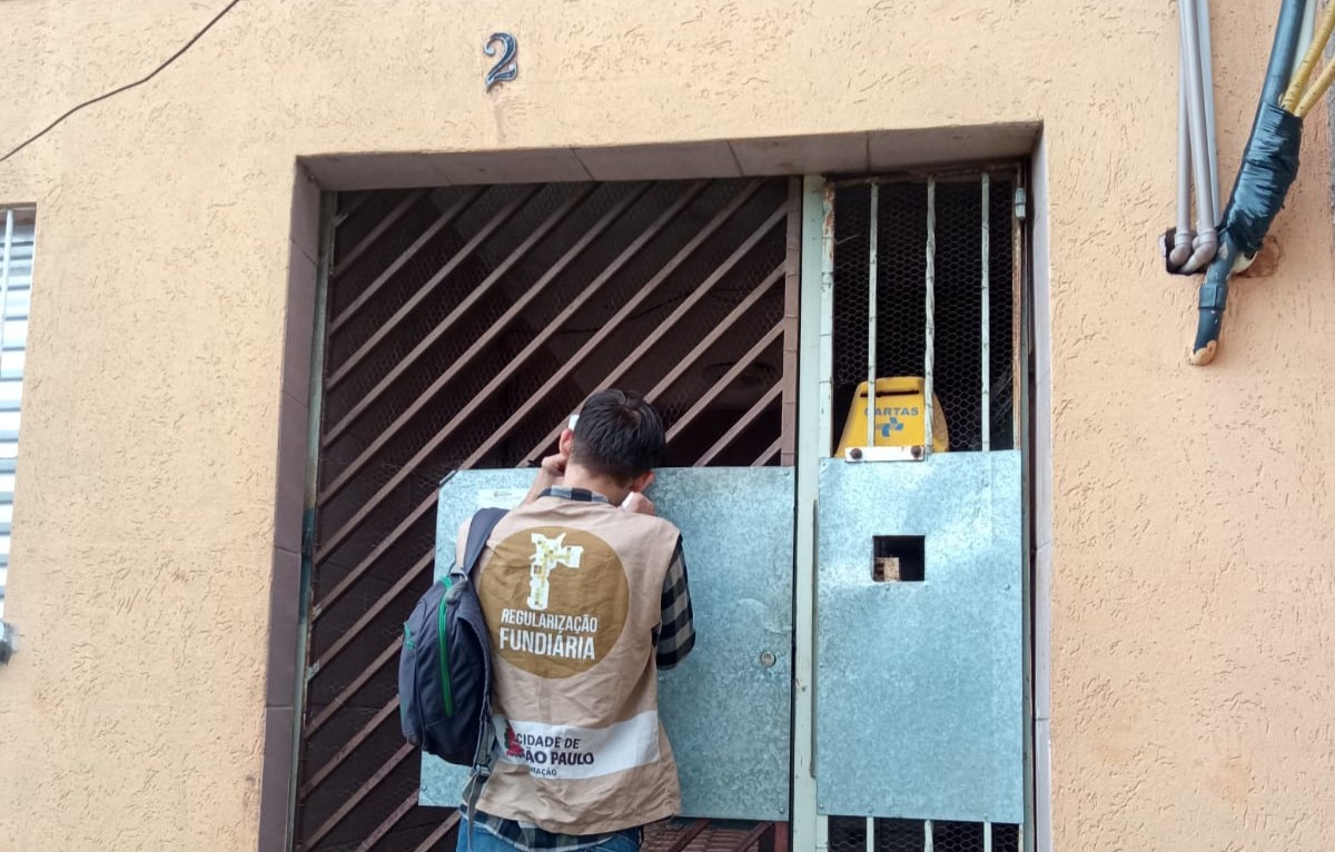 Homem de costas, usando colete bege da Regularização Fundiária da Prefeitura de São Paulo e mochila, conversa pelo interfone de um portão metálico azul em frente a uma casa simples de parede laranja.