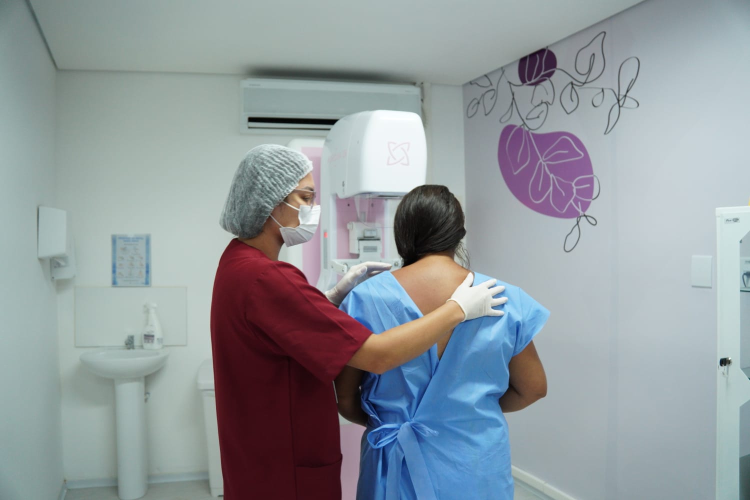 A imagem mostra o interior de uma sala de exame médico, onde uma mulher realiza mamografia. No centro da cena, há duas pessoas vistas de costas. Uma delas é uma profissional de saúde vestindo uniforme vermelho, máscara facial, luvas e uma touca descartável cobrindo o cabelo. Ela está posicionada ao lado de uma paciente e coloca uma das mãos de forma orientadora nas costas dela, ajudando-a a se posicionar corretamente para o exame. A paciente usa uma roupa hospitalar azul aberta nas costas, amarrada na parte inferior, e está voltada para o equipamento de mamografia à sua frente. O aparelho é branco, com uma área interna de cor rosa, onde o exame é realizado. A sala é clara, com paredes brancas e aparência limpa e organizada. No lado esquerdo da imagem há uma pia com torneira e um dispensador de sabão ou álcool. Do lado direito, há um desenho decorativo na parede com linhas que formam folhas e flores em preto e roxo. A cena transmite um momento de preparação para um exame de diagnóstico médico, com a profissional auxiliando a paciente de forma cuidadosa.