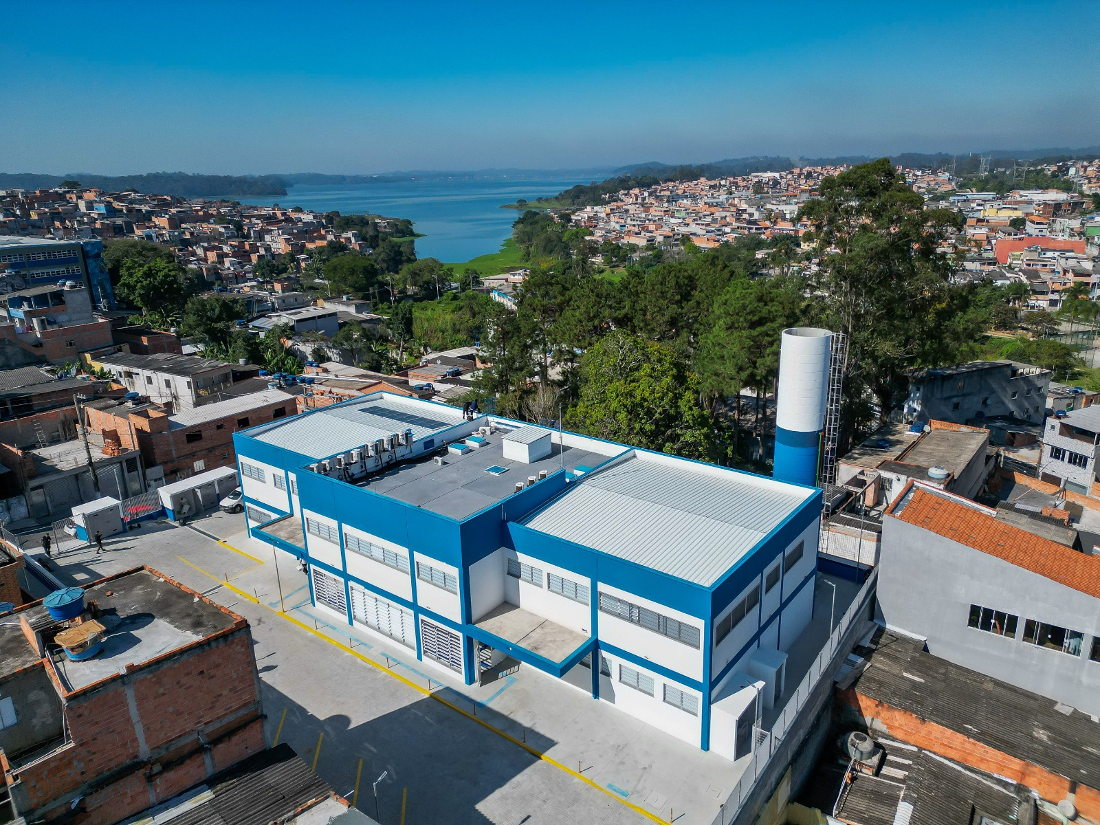 A foto mostra a vista aérea de uma área urbana densamente ocupada, com destaque para um grande edifício de dois andares nas cores branca e azul, localizado em primeiro plano que é a UBS Cantinho do Céu. A construção possui arquitetura moderna, telhado metálico claro, amplas janelas e uma área pavimentada ao redor, sugerindo tratar-se de um equipamento público, institucional ou industrial. Ao redor do prédio, há diversas residências e edificações de alvenaria, distribuídas de forma compacta em uma região de relevo suave. À direita da construção principal, observa-se uma estrutura cilíndrica alta, pintada de azul e branco. Ao fundo da imagem, destaca-se uma extensa represa ou lago de águas calmas, cercada por áreas verdes e bairros residenciais que se estendem pelas margens.