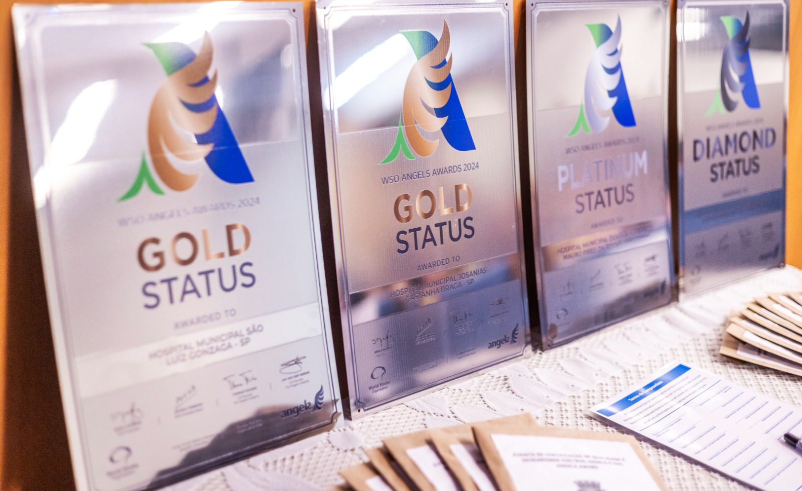 A imagem mostra uma mesa com várias placas de premiação alinhadas lado a lado. Cada placa exibe o logotipo colorido do WSO Angels Awards 2024 e diferentes categorias de reconhecimento, como Gold Status, Platinum Status e Diamond Status. É possível ler que algumas premiações foram concedidas a hospitais municipais de São Paulo. As placas têm acabamento metálico e refletem a luz do ambiente. Sobre a mesa, coberta com uma toalha clara, também há envelopes e um folheto impresso, sugerindo que o material faz parte da organização de um evento de premiação ou cerimônia oficial.