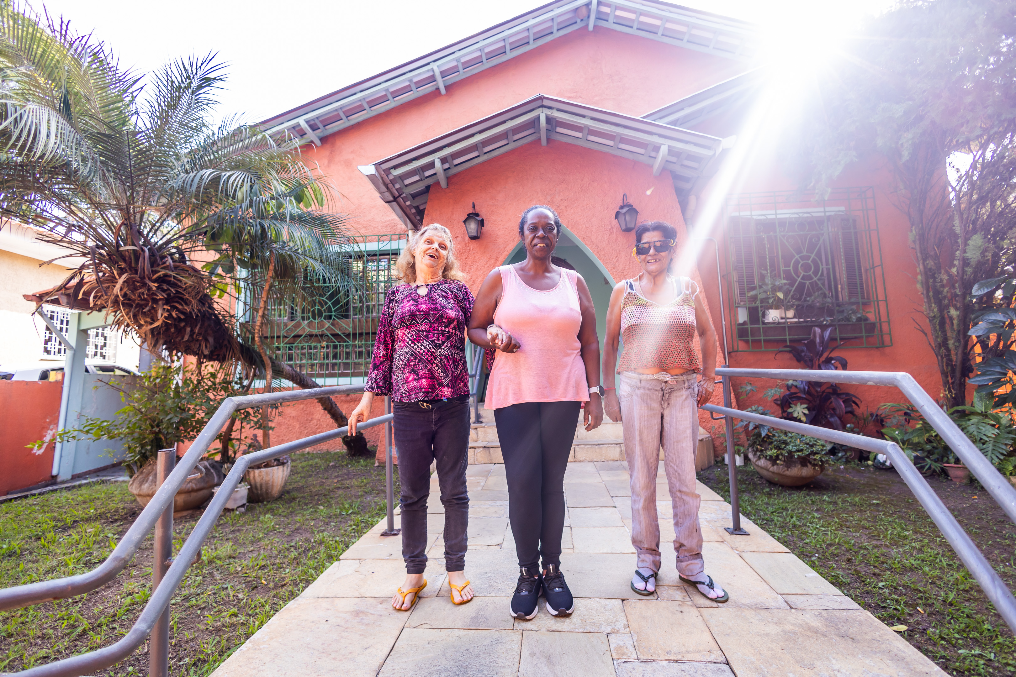 A imagem mostra três mulheres idosas sorrindo e de mãos dadas, em frente a uma residência de cor laranja com arquitetura acolhedora. Elas estão em uma rampa de acesso, ladeada por corrimãos, cercadas por vegetação e plantas ornamentais.  As mulheres aparentam estar bem-dispostas e felizes, e a luz do sol entra pela lateral superior direita da imagem, criando um raio de luz suave que ilumina a cena, dando uma sensação de acolhimento, dignidade e vitalidade. A casa ao fundo tem janelas com grades decoradas, telhado inclinado e uma entrada em arco, transmitindo uma atmosfera de lar.  A imagem sugere um ambiente de residência terapêutica ou institucional acolhedora, voltado para o cuidado e convivência de pessoas idosas em um espaço seguro e humanizado.