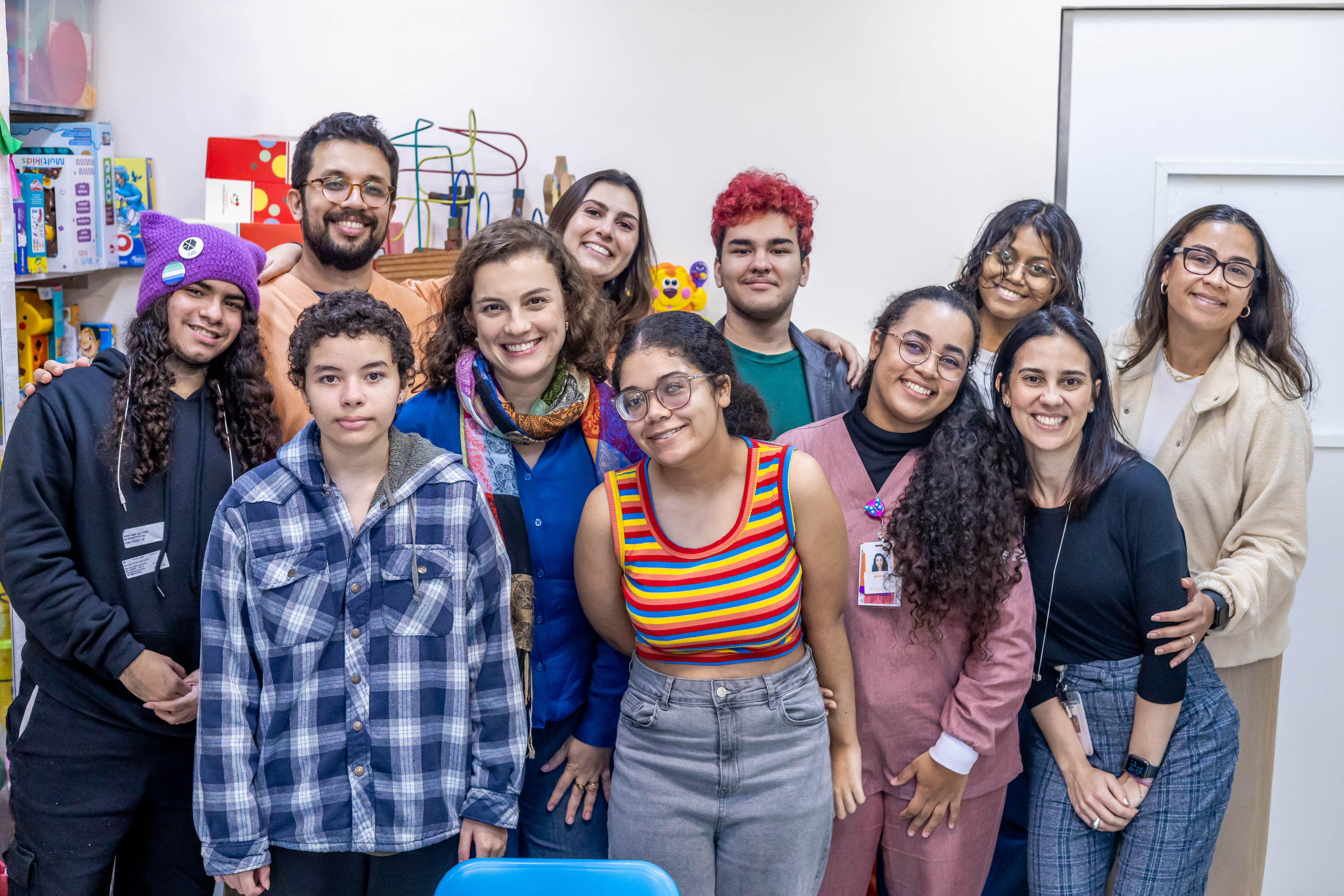 A imagem mostra um grupo de 12 pessoas no Hospital Menino Jesus. Todos estão posicionados próximos uns dos outros e sorrindo para a câmera, transmitindo um clima de acolhimento e amizade.  Na composição há diversidade de idades, estilos e tons de pele. À esquerda, há dois jovens com cabelos longos e cacheados: um deles usa touca roxa com orelhas e moletom preto, e o outro veste uma camisa xadrez azul e cinza. Ao centro, uma mulher adulta de cabelos castanhos curtos e cacheados usa camisa azul e um lenço colorido no pescoço. À frente dela, uma jovem de óculos e cabelos presos está com uma blusa regata listrada em cores vibrantes (vermelho, amarelo, verde, azul), e calça jeans de cintura alta.  Atrás, há uma jovem sorridente com cabelos castanhos lisos e blusa branca, ao lado de um rapaz com cabelo vermelho vibrante, camiseta verde e jaqueta escura. À direita da imagem, estão mais cinco mulheres: uma jovem de jaleco rosa e crachá, de cabelos cacheados e óculos; uma mulher de cabelos lisos pretos, usando blusa preta e calça xadrez azul; uma mulher de óculos, blusa branca e casaco bege; uma moça com cabelo escuro ondulado e blusa clara; e um homem com barba e óculos, vestindo camiseta laranja.