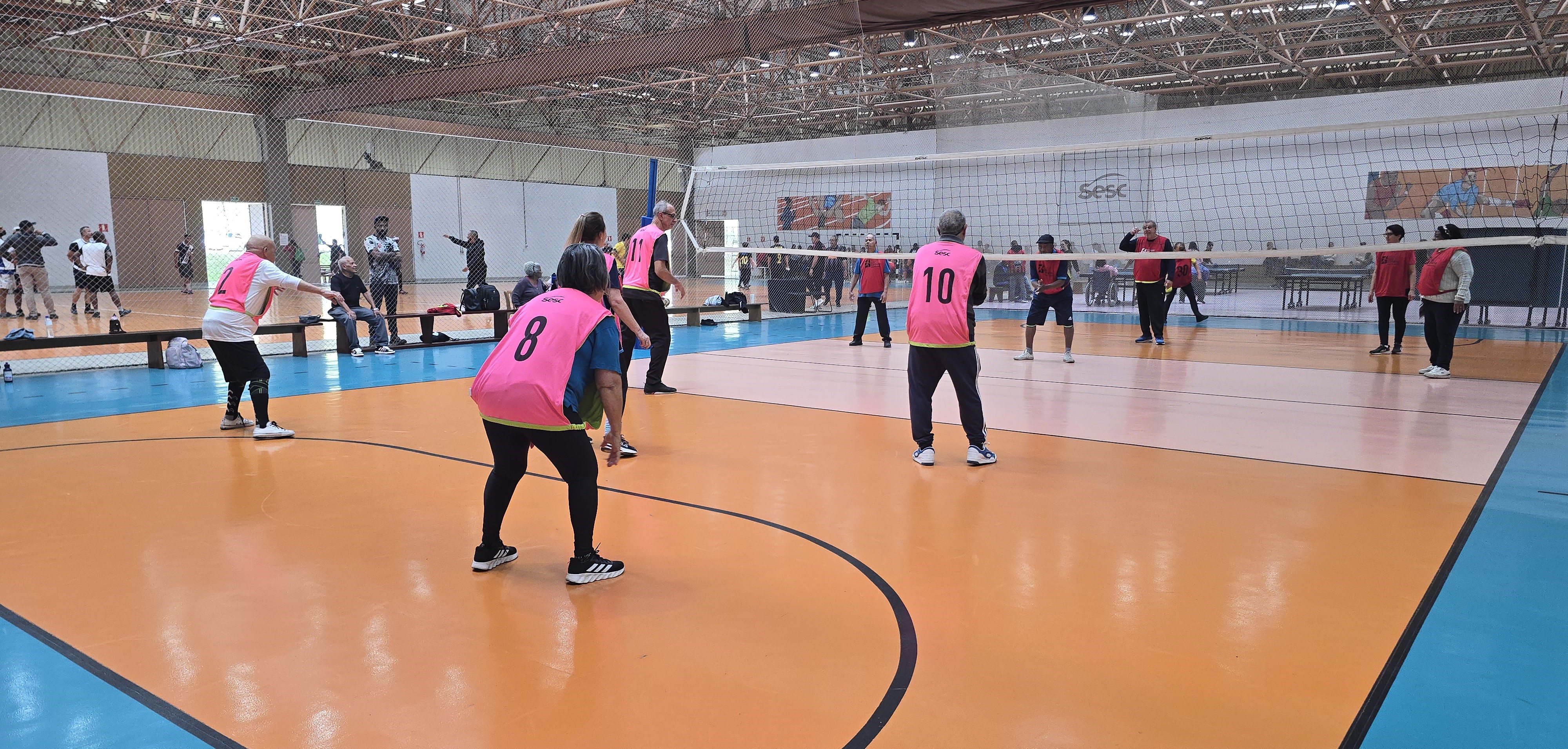 A imagem mostra um grupo de pessoas jogando vôlei adaptado em uma quadra coberta. Os jogadores vestem coletes coloridos: o time em primeiro plano usa coletes rosa com números grandes (2, 8, 10 e 11 visíveis), enquanto o time adversário, do outro lado da rede, usa coletes vermelhos. A quadra tem piso laranja e azul, e ao fundo há um grande painel escrito “SESC”, indicando que a atividade acontece em uma unidade dessa instituição. Algumas pessoas assistem sentadas em bancos ou em pé, do lado de fora da quadra.