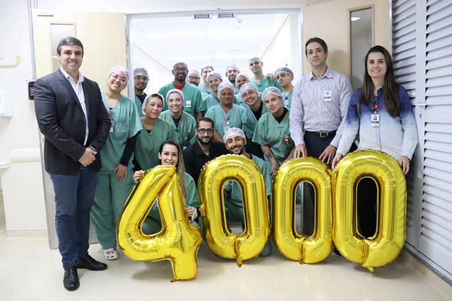 A imagem mostra um grupo de cerca de 15 profissionais de saúde posa em um corredor hospitalar, a maioria vestindo roupas cirúrgicas verdes e toucas. Eles estão sorrindo e reunidos atrás de quatro balões dourados grandes que formam o número “4000”. À esquerda, um homem de terno escuro está em pé com as mãos juntas. À direita, dois profissionais com crachá também participam da foto. O ambiente é iluminado, com paredes claras e portas abertas ao fundo, sugerindo uma área interna do hospital.