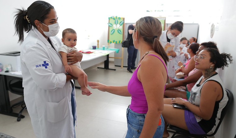 A imagem mostra um atendimento de saúde em uma unidade pública.  Em primeiro plano, uma profissional da área da saúde, vestindo jaleco branco com o símbolo do SUS e usando máscara, segura um bebê no colo. À frente dela, uma mulher, possivelmente a mãe, conversa ou recebe orientações, com a mão estendida em direção à criança.  Ao fundo, há várias mulheres sentadas em cadeiras, muitas com bebês ou crianças, aguardando atendimento. Algumas profissionais também aparecem usando jaleco e segurando pranchetas, indicando organização de fila ou registro de informações.  O ambiente é claro, organizado e parece ser uma sala de atendimento ou vacinação em uma unidade básica de saúde. A cena sugere um momento de cuidado materno-infantil, possivelmente relacionado à vacinação, consulta pediátrica ou acompanhamento de rotina.