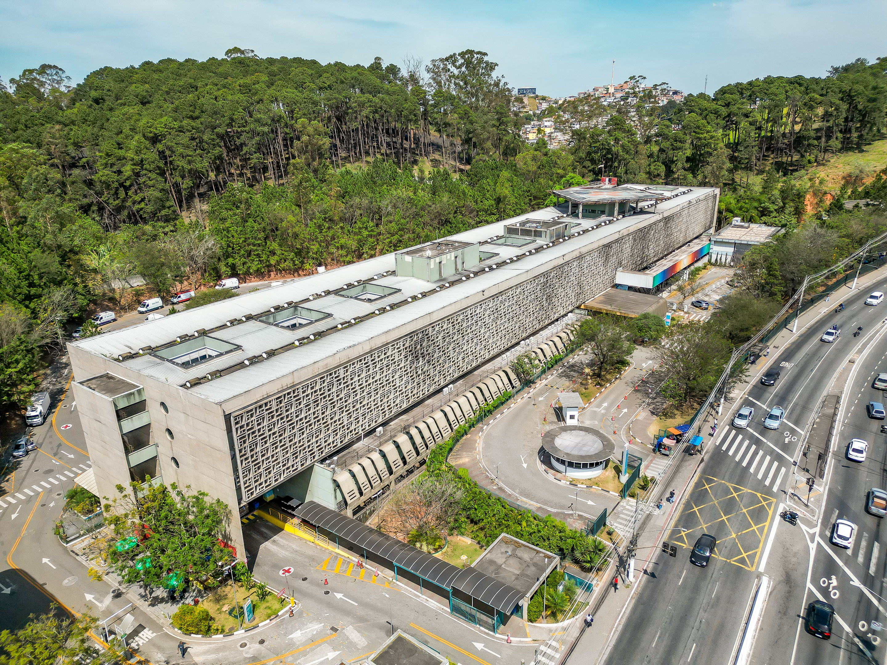 A imagem mostra uma vista aérea do Hospital Municipal Dr. Moyséas Deutsch, localizado em M'Boi Mirim, São Paulo. O edifício é de arquitetura moderna, com uma fachada em concreto e um padrão de janelas no lado esquerdo da construção. Na parte inferior, é possível observar a entrada principal com uma área de estacionamento. Ao redor do hospital, há um grande espaço verde, com árvores de médio porte, sugerindo uma proximidade com a natureza. A rua ao redor do hospital está movimentada, com carros e pedestres, e há faixas de pedestres e sinalização de trânsito visíveis. O edifício está em uma área de cidade, mas com vegetação ao fundo, indicando que o local é cercado por áreas de vegetação e pequenas colinas.