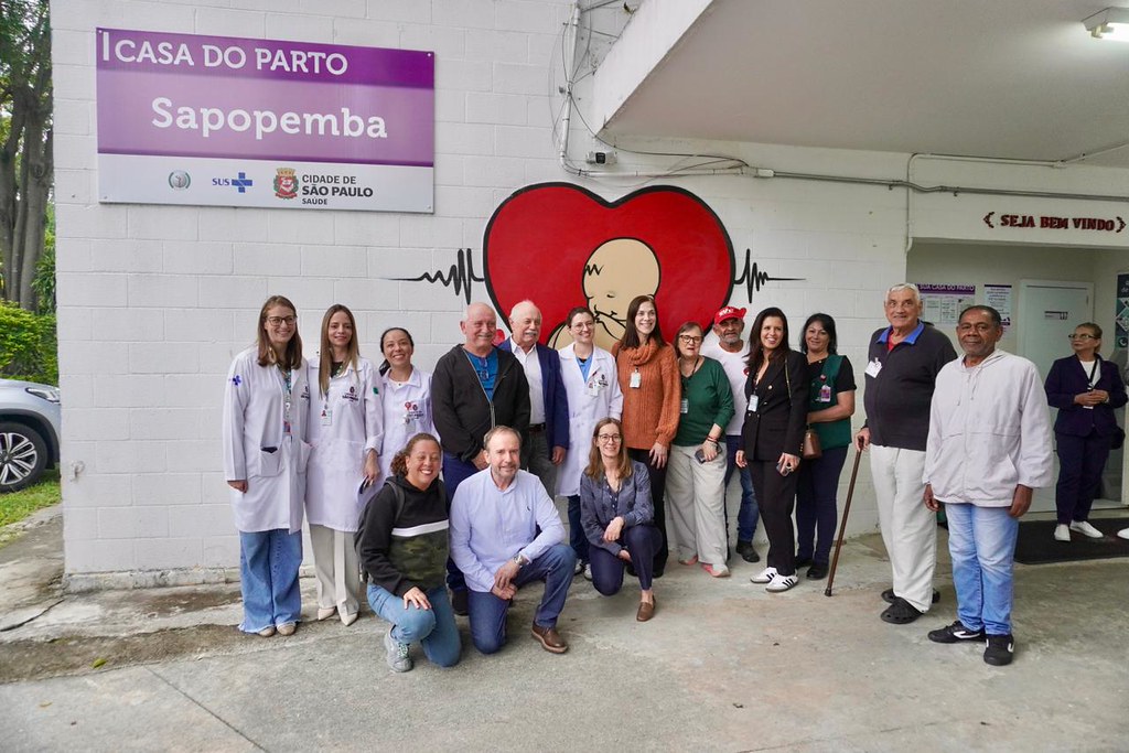 A imagem mostra um grupo de profissionais da saúde em frente à Casa de Parto Sapopemba. Algumas pessoas estão em pé e outras pessoas estão agachadas na frente. Ao centro, há um grande coração vermelho com a imagem de um recém-nascido.