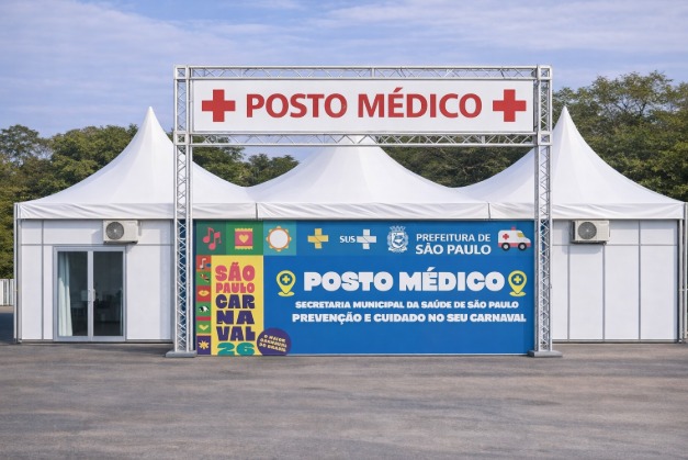 A imagem mostra um posto médico temporário montado ao ar livre, estruturado com tendas brancas, provavelmente para atendimento em um evento público.  Na parte superior, há uma placa grande com uma cruz vermelha e a inscrição “POSTO MÉDICO”, indicando claramente que se trata de um local de primeiros socorros e atendimento de saúde.  Na fachada central, há um painel colorido com várias informações institucionais. Nele aparecem os logotipos do SUS e da Prefeitura de São Paulo, além do texto:  “Posto Médico – Secretaria Municipal da Saúde de São Paulo – Prevenção e Cuidado no seu Carnaval”