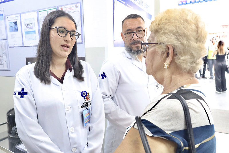 A imagem registra profissionais de saúde em um diálogo próximo e atento com uma paciente idosa. Os profissionais utilizam jalecos brancos com o logotipo oficial da 
