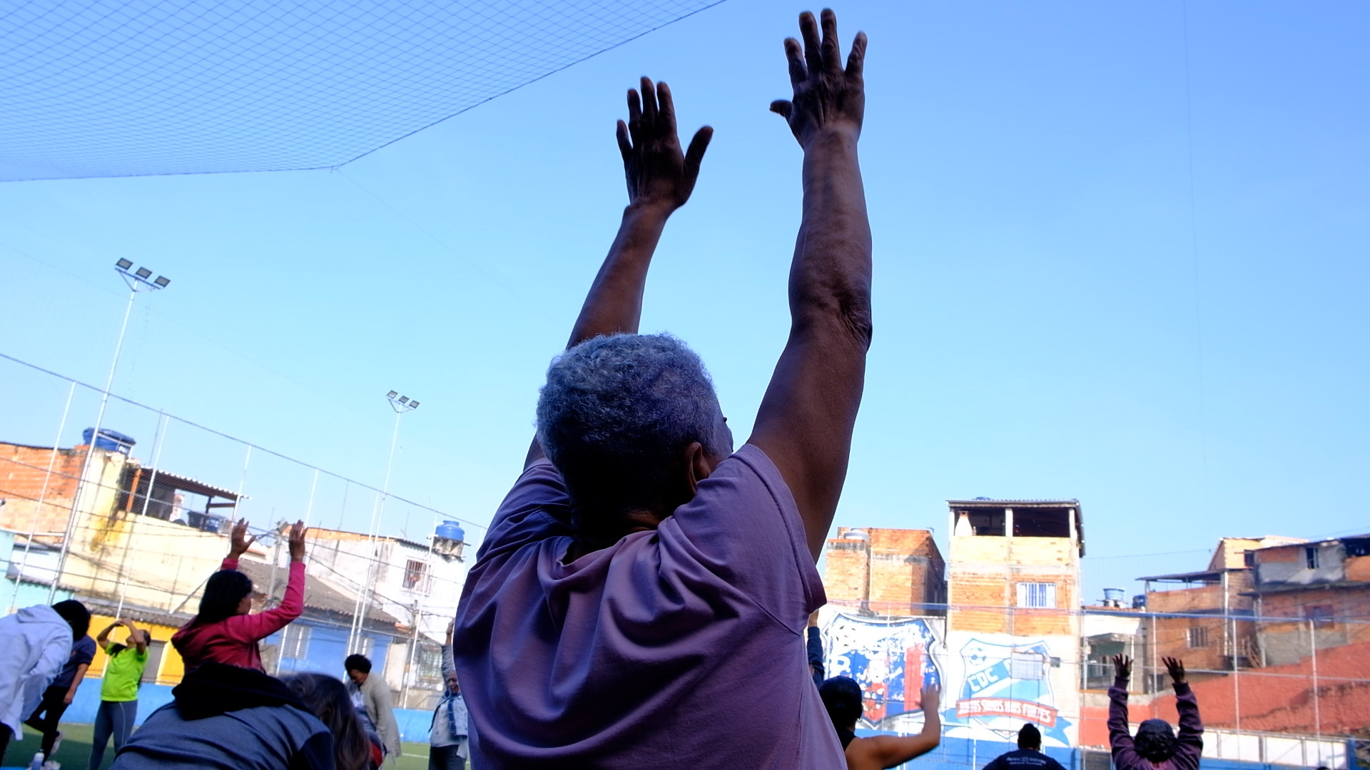 articipantes de uma atividade coletiva das Práticas Integrativas e Complementares em Saúde (Pics) realizam exercícios de alongamento ao ar livre, em uma quadra comunitária da capital. Em primeiro plano, uma mulher aparece de costas com os braços erguidos, acompanhando os movimentos conduzidos em grupo, em uma ação voltada à promoção da saúde, bem-estar e convivência.