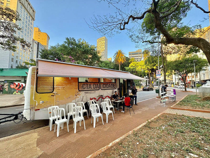A imagem mostra uma unidade móvel de atendimento em saúde instalada em uma calçada arborizada, em uma área urbana. Trata-se de um trailer identificado como “CTA da Cidade”, utilizado para ações de prevenção, testagem e orientação em saúde.  Em frente ao veículo, há várias cadeiras plásticas brancas organizadas em fileiras, indicando que o local recebe atendimento ao público. Algumas pessoas estão sentadas e sendo atendidas por profissionais, enquanto outras aguardam.  O cenário ao redor revela um ambiente típico de centro urbano, com prédios altos ao fundo, árvores, bancos de praça e calçadas largas. À direita, há um pequeno espaço verde com bancos, sugerindo que a ação ocorre em uma praça ou área pública.  A iluminação natural e o céu azul indicam que a foto foi feita durante o dia, em horário de boa luminosidade. A composição transmite a ideia de um serviço de saúde acessível, realizado fora das unidades tradicionais, diretamente no espaço público, aproximando o atendimento da população