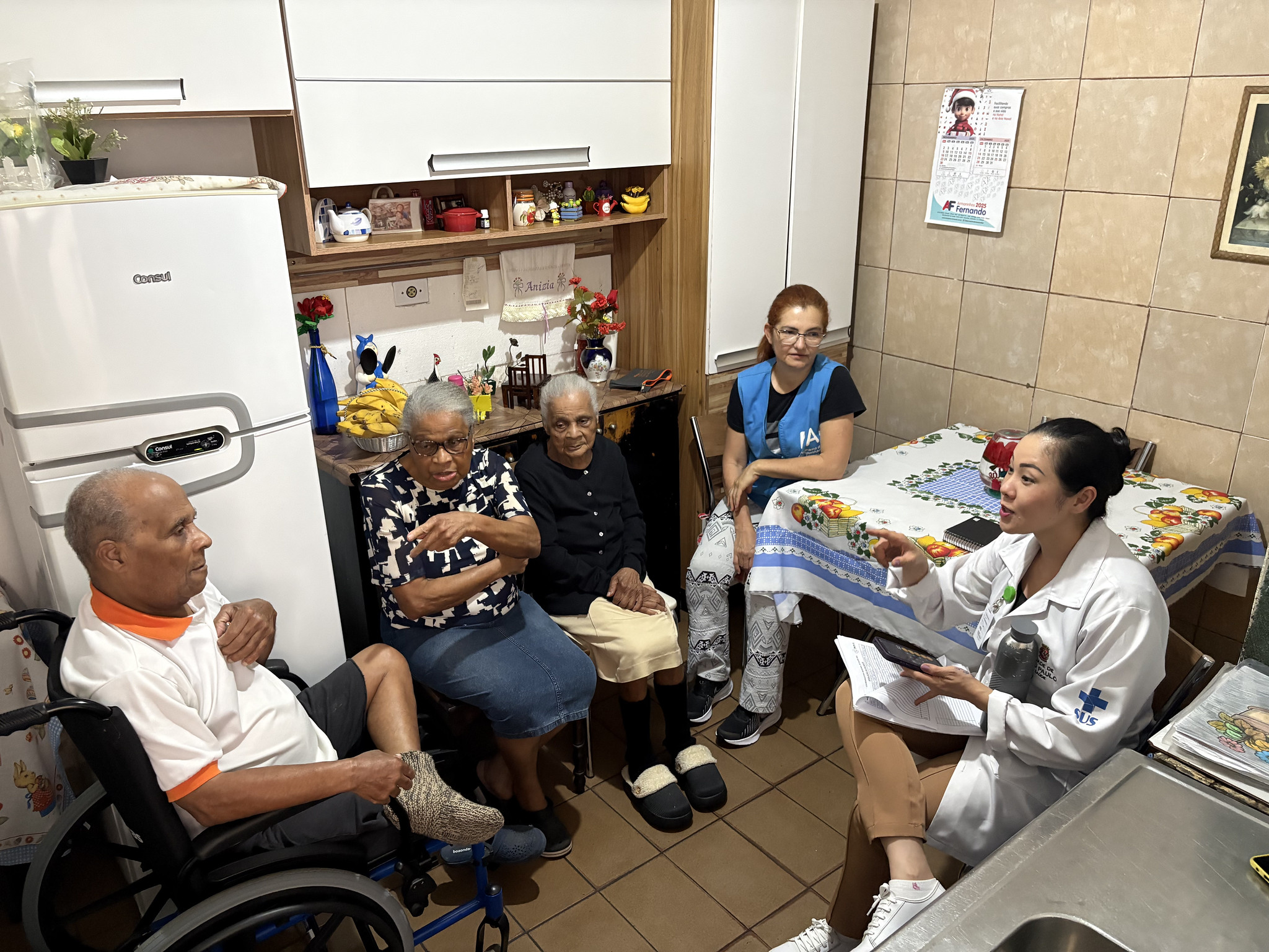 A foto registra um atendimento de saúde domiciliar em uma cozinha residencial simples e iluminada. Cinco pessoas estão no ambiente: três idosos (dois sentados e um em cadeira de rodas) e duas profissionais de saúde do Programa de Acompanhante de Idosos. O grupo está organizado em um semicírculo, sugerindo uma conversa atenciosa. As Pessoas (da esquerda para a direita) Homem Idoso: Está sentado em uma cadeira de rodas manual, de perfil para a direita. Ele é negro, tem cabelos curtos grisalhos e usa uma camisa polo branca com gola laranja e bermuda escura. Ele ouve atentamente o que a profissional de saúde diz. Mulher Idosa (ao centro): Está sentada em uma cadeira comum. Ela é negra, usa óculos de grau e uma blusa estampada em preto e branco. Ela aponta com o dedo indicador em direção à profissional, como se estivesse explicando algo. Mulher Idosa (à direita do centro): Uma senhora negra, com cabelos brancos presos, usando uma blusa preta de manga longa, saia bege e pantufas pretas. Ela observa a cena de forma serena. Agente de Saúde: Uma mulher de pele clara e cabelos ruivos amarrados, sentada ao fundo, entre as idosas e a mesa. Ela veste uma camiseta preta sob um colete azul (uniforme de agente de saúde) e calça estampada. Profissional de Saúde (Médica ou Enfermeira): Sentada à direita, em primeiro plano e de perfil para a esquerda. Ela é uma mulher jovem de traços orientais, com o cabelo preso em um coque. Veste um jaleco branco com o logotipo do SUS (Sistema Único de Saúde) no braço. Ela segura um celular e papéis, gesticulando com a mão direita enquanto fala com os idosos.