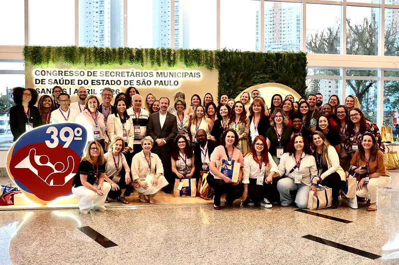 Esta imagem registra um grande grupo de pessoas reunidas em um evento institucional. O cenário é um saguão amplo e iluminado, com grandes janelas de vidro ao fundo que mostram edifícios urbanos. No centro superior, um painel decorado com folhagens traz o título: