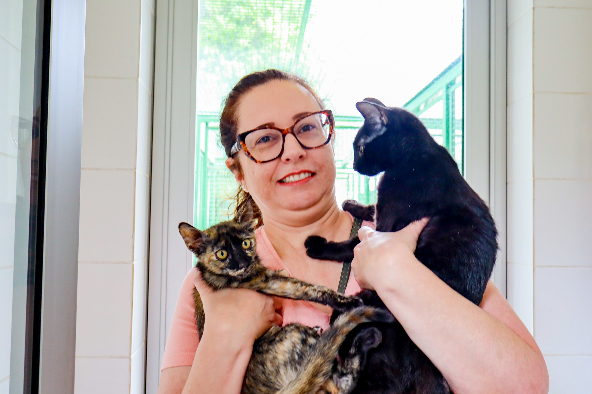 Uma mulher segura dois gatos no colo dentro de um ambiente claro, próximo a uma janela. Ela sorri enquanto apoia um gato preto com uma das mãos e um gato de pelagem mesclada (preta, marrom e bege) com a outra. Os animais parecem atentos e tranquilos, voltados em direções diferentes. Ao fundo, é possível ver uma área externa cercada, sugerindo um espaço de acolhimento e cuidado para os animais.