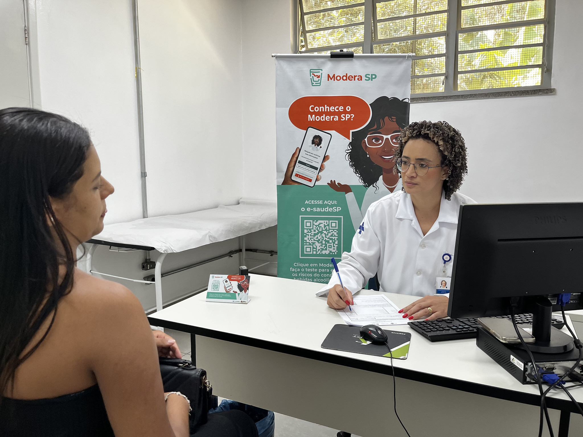Profissional de saúde realiza atendimento em consultório de uma Unidade Básica de Saúde (UBS), durante orientação sobre a ferramenta Modera SP. Ao fundo, um banner do programa apresenta informações sobre o acesso à plataforma pelo aplicativo e-saúdeSP, com QR Code para consulta e avaliação dos riscos relacionados ao consumo de bebidas alcoólicas.